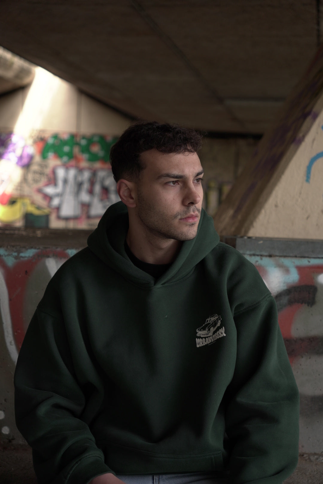 Modelo con Hoodie Urban Essentials verde frente a graffiti urbano - sudadera streetwear con capucha UVX Studio